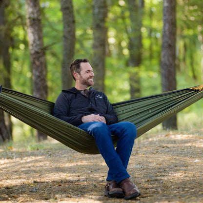 Man relaxing in a green Eclipse Heated Hammock Tent set up among trees, showcasing its portability and comfort for camping.