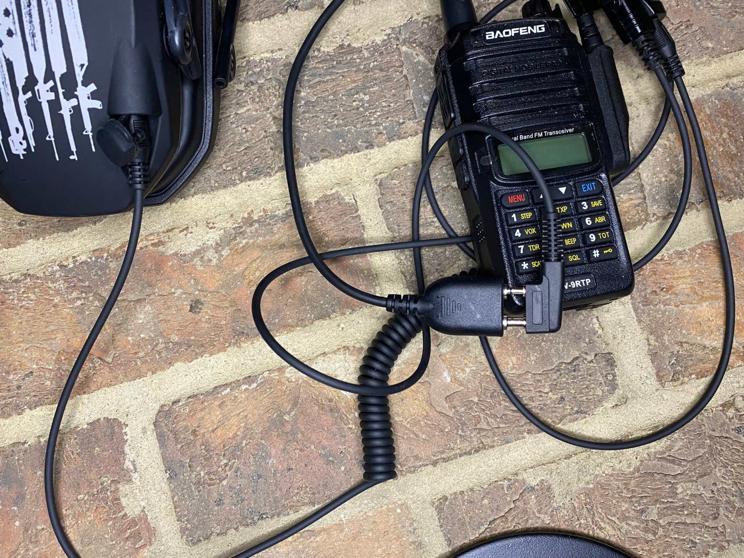 SMC Baofeng UV9R, GT-3WP cable connecting radio to earmuffs on brick background.