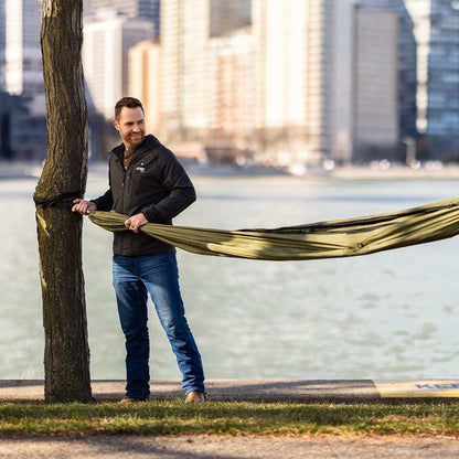 Man setting up Eclipse Heated Hammock Tent in Pine color by waterfront, showcasing portable setup and outdoor use.