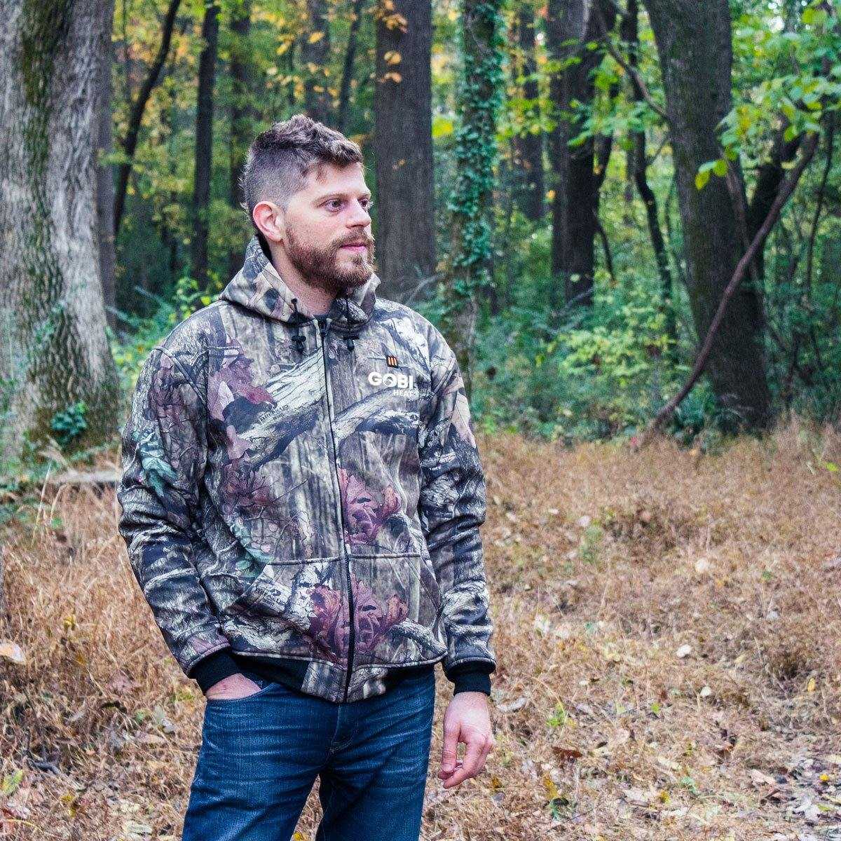 Man wearing Shadow Heated Hunting Hoodie in Mossy Oak Camo pattern standing in forest.