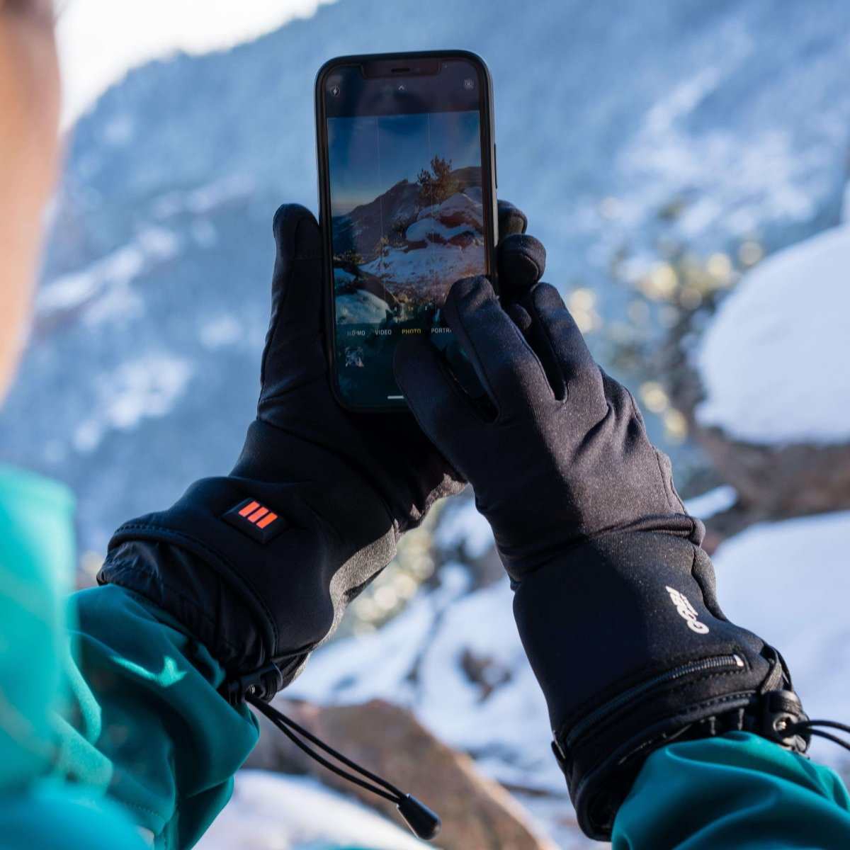 Person using touchscreen device with Stealth Heated Glove Liners in snowy outdoor setting.