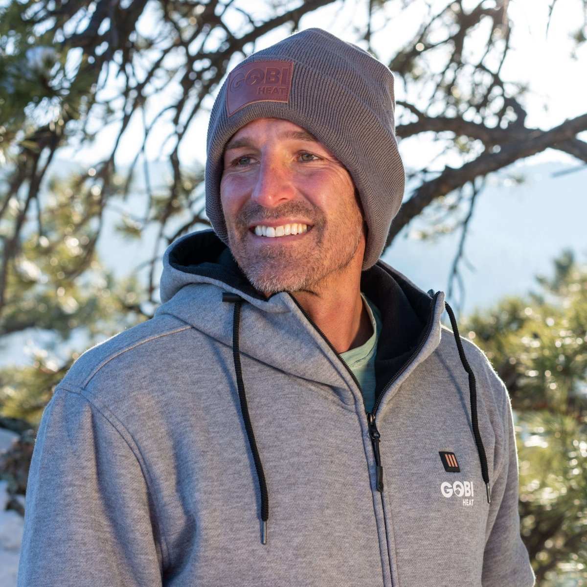 Man wearing Summit Mens Heated Beanie in gray, outdoors in winter with trees in the background.