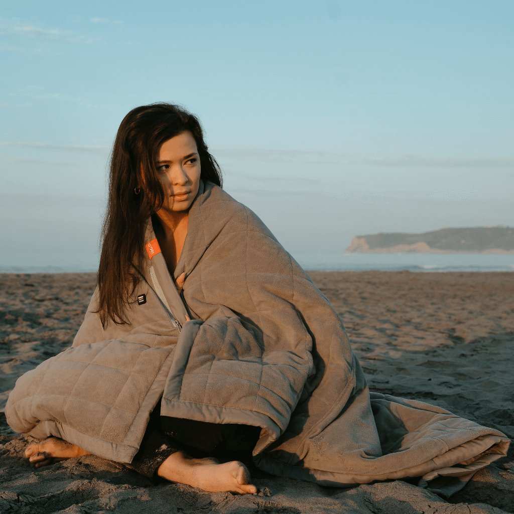 Woman sitting on beach wrapped in Zen Portable Heated Blanket.