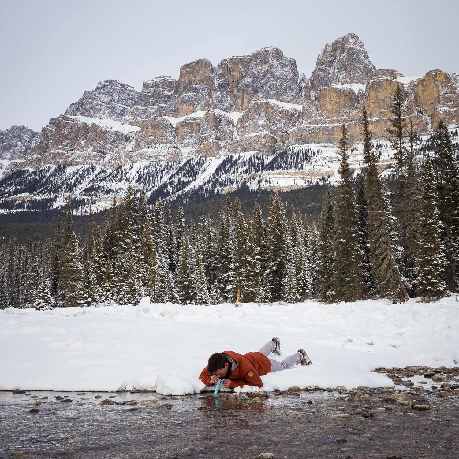 Person using LifeStraw Personal Water Filter to drink from a stream in snowy mountain landscape.